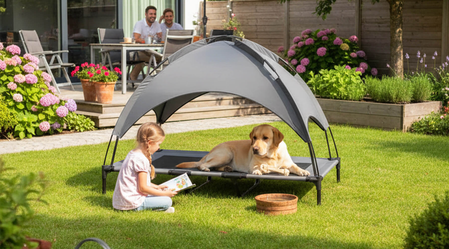 Ein heller Hund entspannt auf einer erhöhten Hundeliege mit Sonnendach im grünen Garten, während ein Kind daneben im Gras liest – perfekter Liegeplatz für den Sommer.