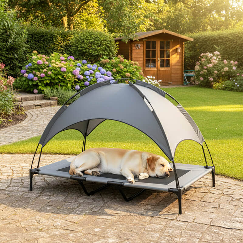 Ein heller Labrador Retriever schläft entspannt auf einer erhöhten Outdoor Hundeliege mit grauem Sonnendach im Garten. Im Hintergrund sind blühende Hortensien, eine Rasenfläche und ein Holzgartenhaus zu sehen.