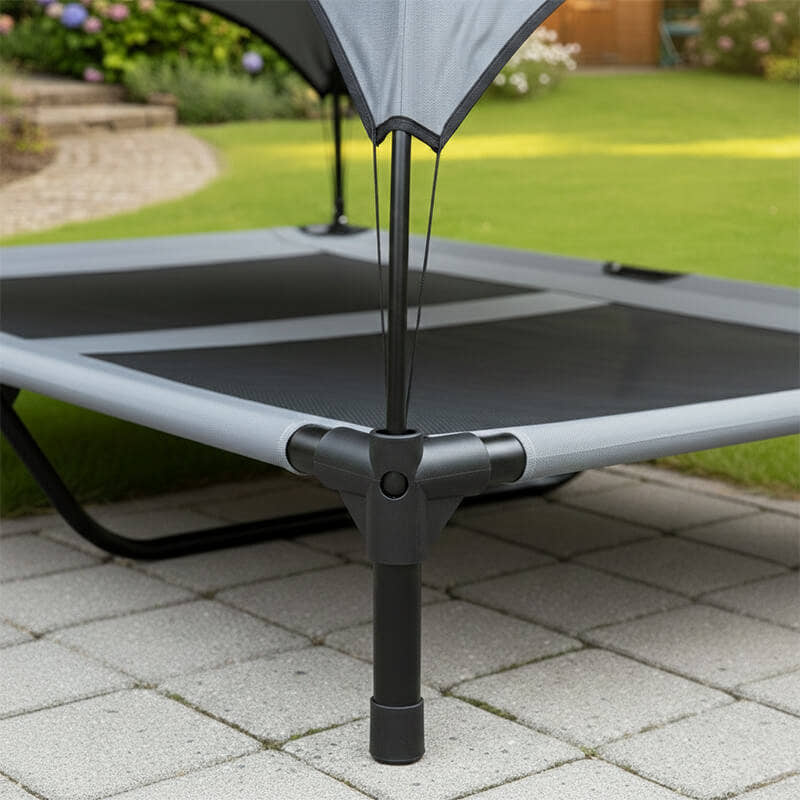Nahaufnahme der robusten Eckverbindung und des stabilen Standfußes einer erhöhten Outdoor Hundeliege. Das Detailbild zeigt das schwarze Metallgestänge, das wetterfeste graue Gewebe und die sichere Steckmontage auf einem gepflasterten Gartenweg.